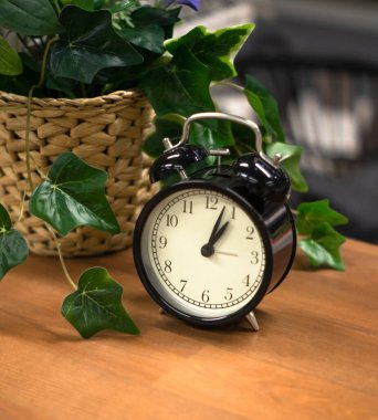 Eski bir çalar saat ahşap bir masanın üzerinde duruyor ve yanında yeşil bitkilerle dolu bir hasır sepet var..