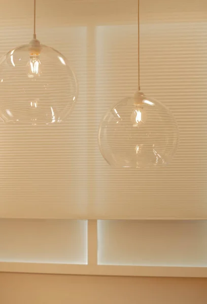 Parlak iç kısımda, tavandan sarkan büyük yuvarlak gölgeli iki cam lamba. Arka plandaki bej tekerlekli jaluzilerde.