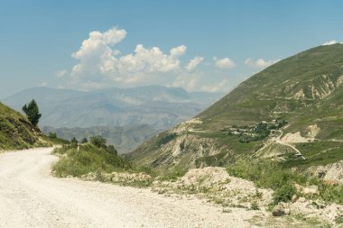 Uzak mesafeden küçük bir dağ köyünün görülebileceği hafif bir dağ toprak yolu. Beyaz bulutlu mavi bir gökyüzünün altında.