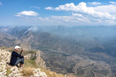 Siyah gömlekli, dar pantolonlu, beyaz şapkalı, kısa saçlı genç bir kadın inanılmaz güzel bir dağ manzarasında kayalık bir uçurumda oturuyor..
