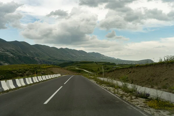 Beyaz şeritli bir asfalt yol, önünde yeşil tepeler ve uzakta yüksek dağlar görülebilir. Gökyüzü beyaz bulutlarla dolu..