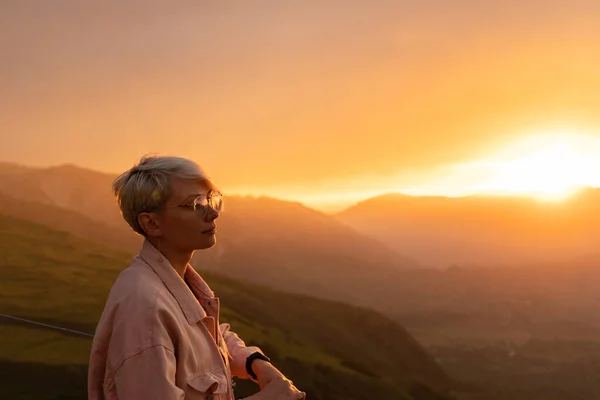Göz kamaştırıcı bir kırmızı ve sarı gün batımının arka planında, genç bir kadın, yakın plan, kısa, beyaz saçlı, şeffaf gözlüklü, soluk pembe kot ceketli, duruyor..