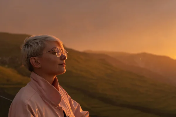 Göz kamaştırıcı bir kırmızı ve sarı gün batımının arka planında, genç bir kadın, yakın plan, kısa, beyaz saçlı, şeffaf gözlüklü, soluk pembe kot ceketli, duruyor..