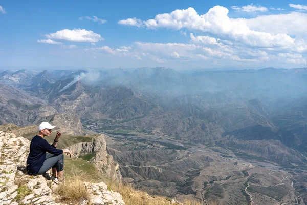 Siyah gömlekli, dar pantolonlu, beyaz şapkalı, kısa saçlı genç bir kadın inanılmaz güzel bir dağ manzarasında kayalık bir uçurumda oturuyor..