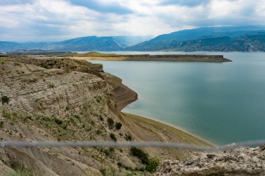 Alışılmadık derecede güzel bir göl, bulutlu mavi gökyüzünün üzerindeki yüksek dağların arasında bir su deposu. Ön planda çiçekler, taşlar, bir kaya var..