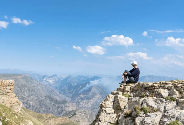 Siyah gömlekli, dar pantolonlu, beyaz şapkalı, kısa saçlı genç bir kadın inanılmaz güzel bir dağ manzarasında kayalık bir uçurumda oturuyor..