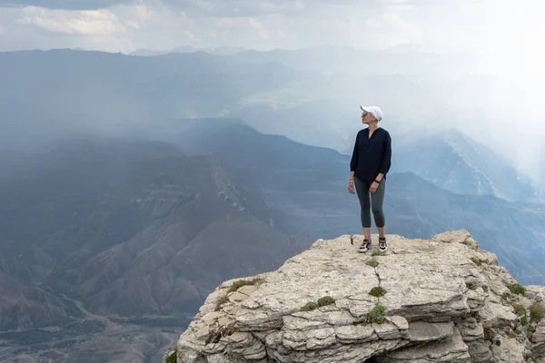 Siyah gömlekli, dar pantolonlu, beyaz şapkalı, kısa saçlı bir kadın inanılmaz güzel bir dağ manzarasında kayalık bir uçurumda duruyor..
