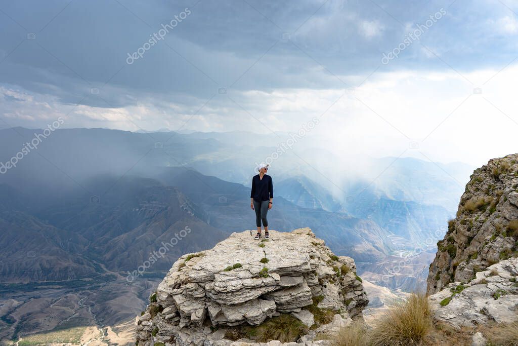 A young woman in a black shirt, tight-fitting trousers, white cap, short hair, stands on a rocky cliff in an incredibly beautiful mountain landscape.