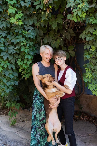 Anne ve genç kız yan yana duruyorlar, sarılıyorlar ve gülüyorlar, kollarında büyük bir köpek tutuyorlar. Okul üniformalı kız..