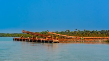 Outdoor industrial jetty at river bank with incline large conveyor for transportation bauxite ore from mining shuttle trains to mini bulk carrier (feeder) ships. Guinea, West Africa. clipart