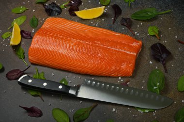 Masadaki taze çiğ somon balığı filetosunu mutfak bıçağı, limon dilimleri ve salata yaprakları ile kapayın.