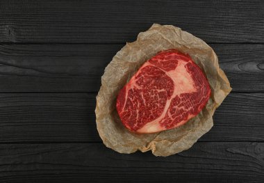 Close up one aged prime marbled raw ribeye beef steak on brown paper parchment wrapping over black wooden table background, elevated top view, directly above