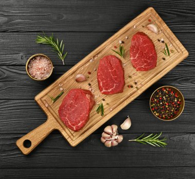 Close up three aged prime marbled raw tenderloin or fillet mignon beef steaks on brown oak wood cutting board, with spices, over black wooden table background, elevated top view, directly above