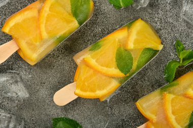 Taze portakal dilimli üç meyveli dondurma, gri masa yüzeyinde yeşil nane yaprakları ve buz küpleri, üst manzaranın hemen üstünde.