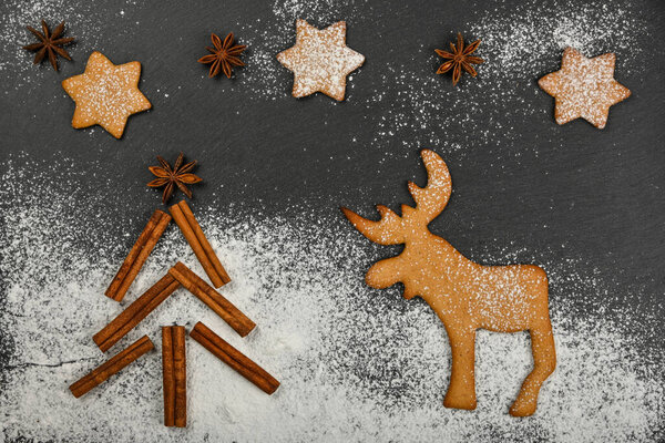 Close up gingerbread cookies with cinnamon Christmas tree, reindeer and star anise on black slate background with powdered white sugar icing and copy space, elevated top view, directly above