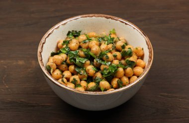 Ceramic bowl of cooked seasoned chickpeas with chopped parsley and onion green herbs rests on a dark brown wooden table, offering a healthy vegan meal option, copy space