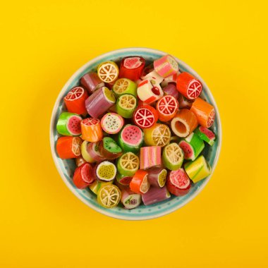 Small pieces of colorful fruit hard sugar candy filling a small bowl placed on a vibrant yellow background with copy space
