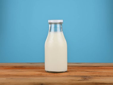 One glass milk bottle closed with white metal cap standing on rustic wooden table with light blue background, representing freshness and healthy lifestyle