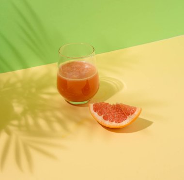 Greyfurt suyu ve sarı ve yeşil arka planda palmiye yapraklarıyla dilimlenmiş bir bardak. Yaz meyveli içecek ve içecek konsepti. Durgun bir bakış açısı.