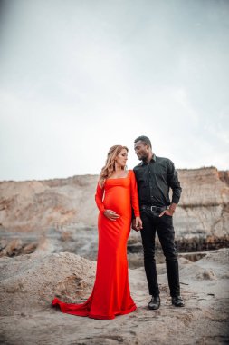 Granit kanyona karşı poz veren çok ırklı çiftin portresi. Şık kırmızı elbise giyen hamile bir kadın. 