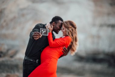  Çok ırklı bir çift granit kanyona karşı ultrason fotoğrafı tutuyor. Şık kırmızı elbise giyen hamile bir kadın. 