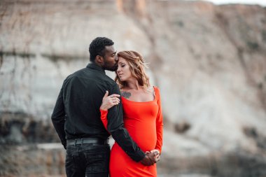 Granit kanyona karşı poz veren çok ırklı çiftin portresi. Şık kırmızı elbise giyen hamile bir kadın. 