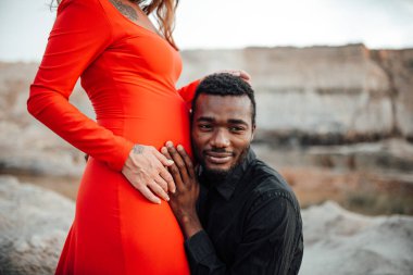 Granit kanyona karşı poz veren çift. Afrikalı bir adam kırmızı elbiseli hamile bir kadının karnına dokunuyor. 