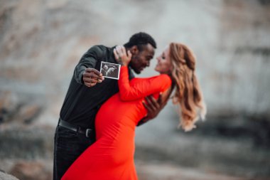  Çok ırklı bir çift granit kanyona karşı ultrason fotoğrafı tutuyor. Şık kırmızı elbise giyen hamile bir kadın. 