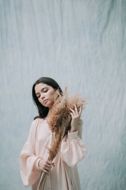 Kurumuş çiçeklerle poz veren esmer kadın. Pembe elbiseli, kumaş arka planda. 