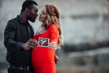  Çok ırklı bir çift granit kanyona karşı ultrason fotoğrafı tutuyor. Şık kırmızı elbise giyen hamile bir kadın. 