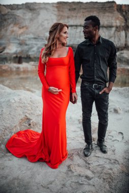 Granit kanyona karşı poz veren çok ırklı çiftin portresi. Şık kırmızı elbise giyen hamile bir kadın. 