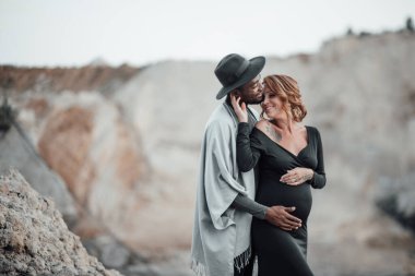 Siyah elbiseli hamile kadını kucaklayan Afrikalı Amerikalı bir adam. Granit kanyona karşı poz veren bir çift.