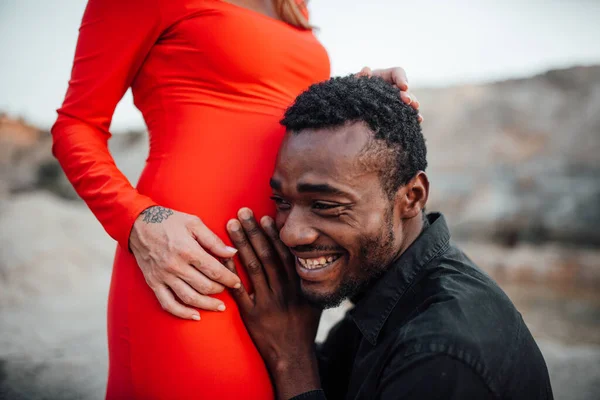 Granit kanyona karşı poz veren çift. Afrikalı bir adam kırmızı elbiseli hamile bir kadının karnına dokunuyor. 