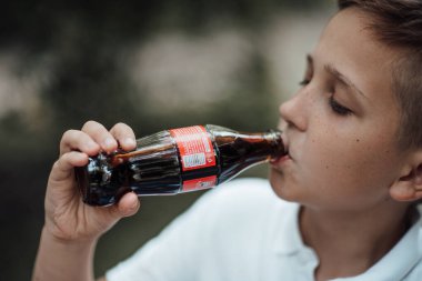 Beyaz tişörtlü, bulanık arka planda kola içen çocuğun yakın çekimi. 