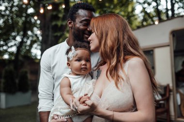 Küçük kızı kucaklayan sevgi dolu bir çiftin açık hava portresi. Aile parkta dinleniyor. Arka planda karavan var.