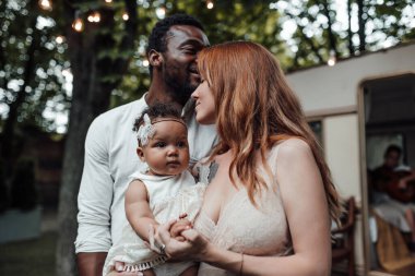 Küçük kızı kucaklayan sevgi dolu bir çiftin açık hava portresi. Aile parkta dinleniyor. Arka planda karavan var.