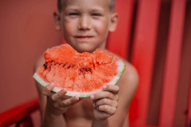 Küçük çocuk kırmızı sandalyede oturuyor ve karpuz yiyor. 