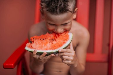 Küçük çocuk kırmızı sandalyede oturuyor ve karpuz yiyor. 