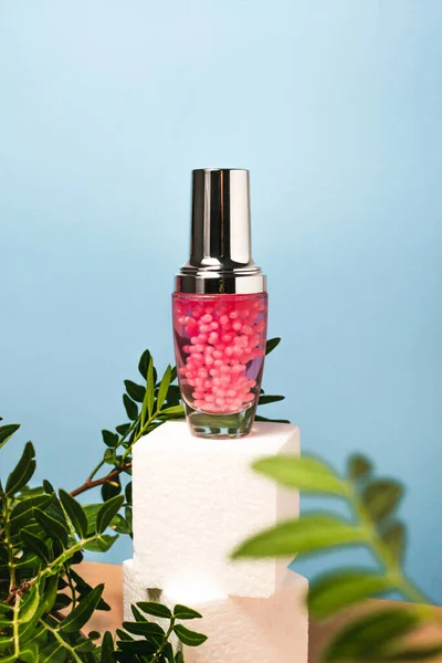 Mavi arka planda pembe granüllü kozmetik serum şişesi. Güzellik sağlığı doğal bir kavramdır. Yaya ürünleri yeşil yapraklı yaratıcı bir konsept sunar. Kopya alanı ile kare küp platformu.
