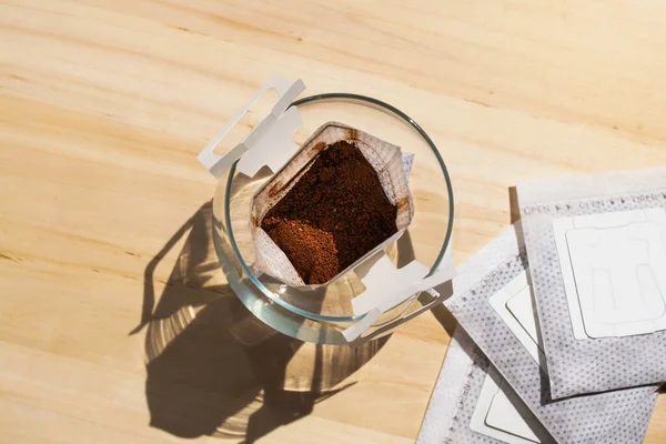 Glass cup with drip coffee bags on wooden table background. Instant paper sachet above still life. Trendy hot beverage home brew. Modern natural roasted aromatic drink. Fresh morning making coffee