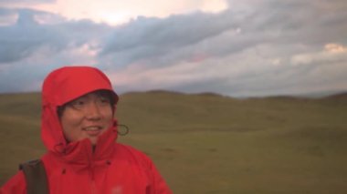 Turuncu ceketli Asyalı adam dağlarda dönüp kamerayla ateş ediyor.