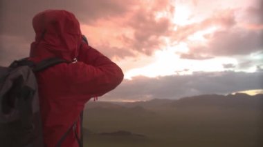 Dışarıdaki turuncu ceketli fotoğrafçı tepenin üstünde gün batımında fotoğraf çekiyor.