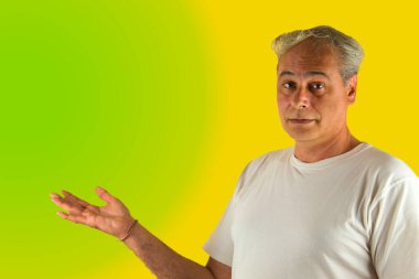 Gri saçlı adam yeşil ve sarı arka planda eli olan bir şey gösteriyor.