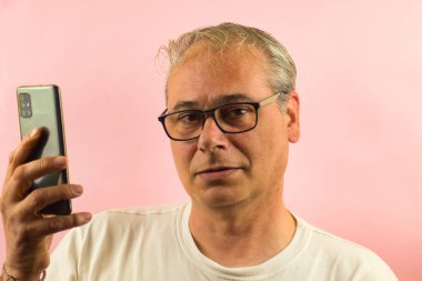 Gri saçlı, gözlüklü, elinde cep telefonu olan bir adamın pembe arka planda önümüze baktığı yakın plan.