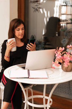 Evde çalışan bir kadın dizüstü bilgisayar kullanıyor telefonuna bakıyor ve elinde bir bardak tutuyor.