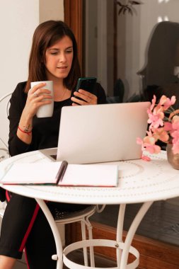 Evde çalışan bir kadın dizüstü bilgisayar kullanıyor telefonuna bakıyor ve elinde bir bardak tutuyor.