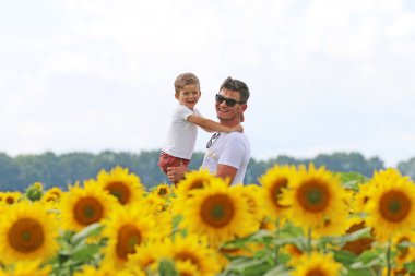 happy joyfull father with his cute clever well mannered  son in field of blossoming sunflower clipart
