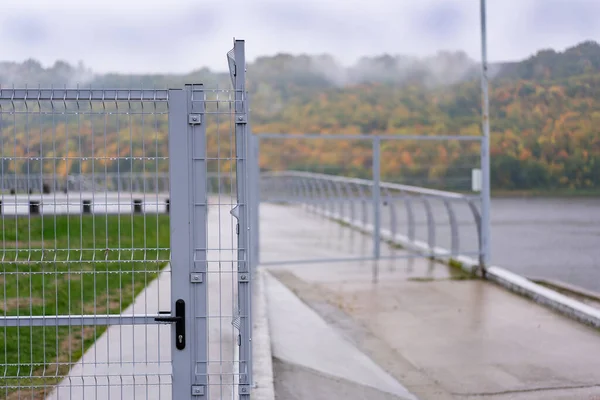The fence is metal lattice with a gate and a lock on the background of the river and the colorful autumn forest. The background is blurred