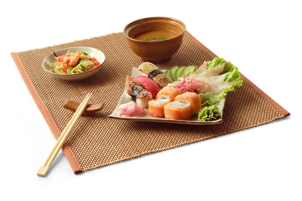 Asiatiska affärslunch med rullar, sashimi, isumi-dai, maguro, unagi, tomat, misosoppa, sallad och pinnar på mattor över vit bakgrund — Stockfoto