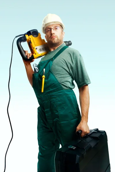 Seriöser Arbeiter mit Helm, Brille und Overall hält Perforator auf der Schulter und großen schwarzen Koffer in der Hand vor weißem Hintergrund — Stockfoto